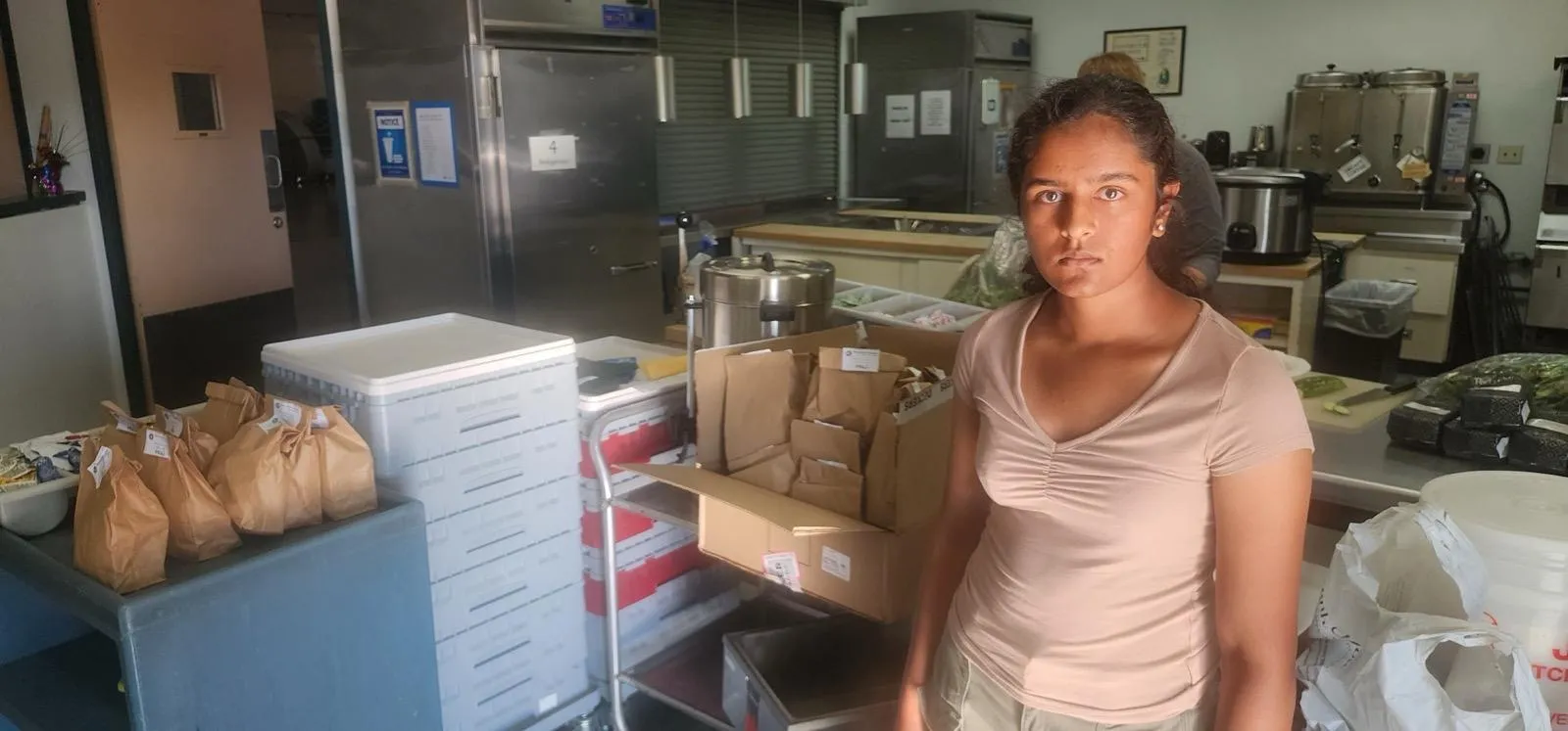 Isha in a commercial kitchen beside a box of packed paper lunch bags, with industrial refrigerators and steam kettles behind her.