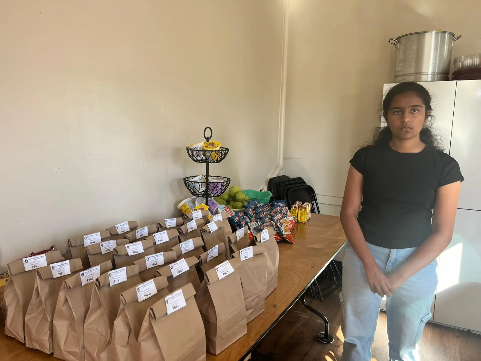 Dozens of labelled paper lunch bags on a table, with chips, apples, fruit cups, and juice boxes staged beside them.