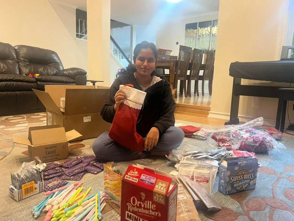 Isha sitting on the floor holding a finished stocking, surrounded by open boxes of supplies.