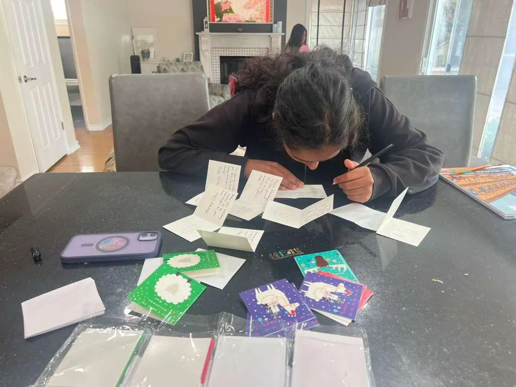 Isha writing holiday cards by hand at a table covered in open cards and sealed packets.
