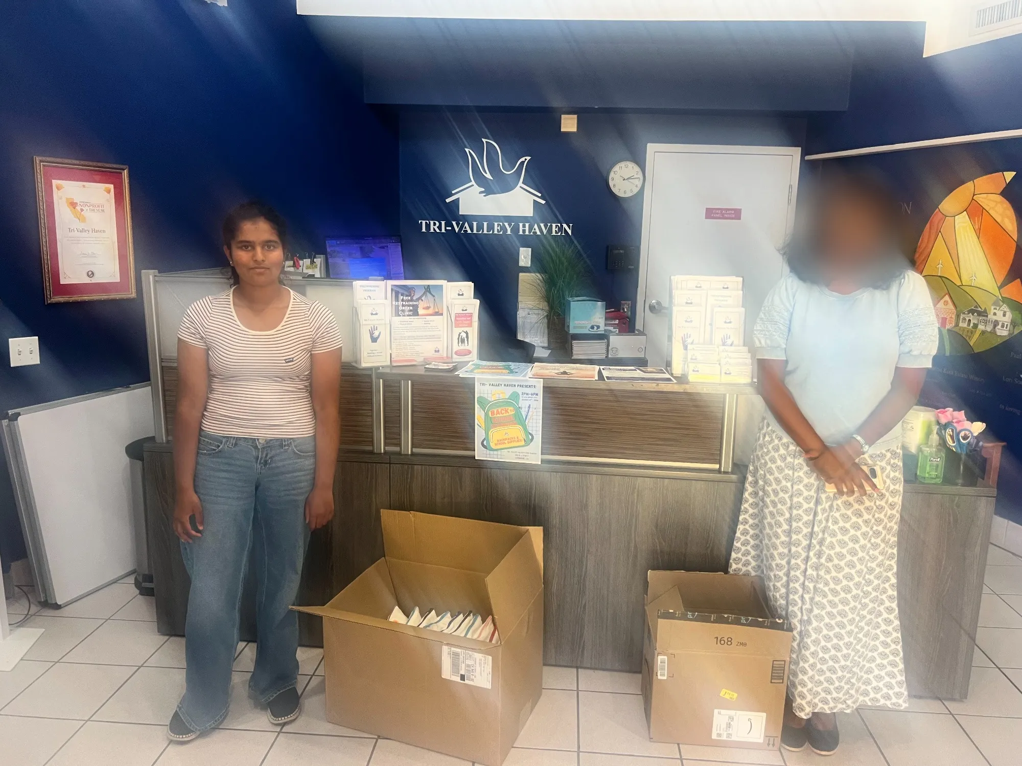 Isha standing beside two boxes of donated supplies in the Tri-Valley Haven lobby.